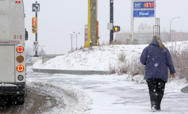 a woman in a blue winter coat walks along a snowy sidewalk as a city bus goes by. 