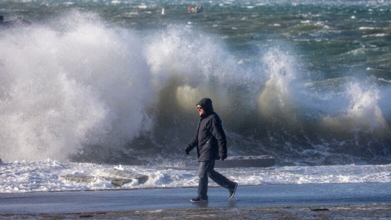 A man walks along the beach with a large wave crashing in the distance behind him. 