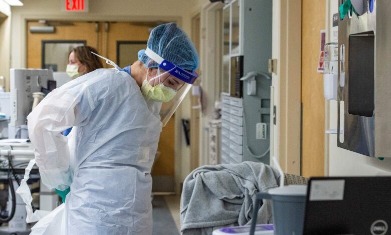 A female health-care worker wearing full personal protective equipment, including a yellow medical mask, face shield and blue latex gloves, does up the ties of her white plastic medical gown behind her back.