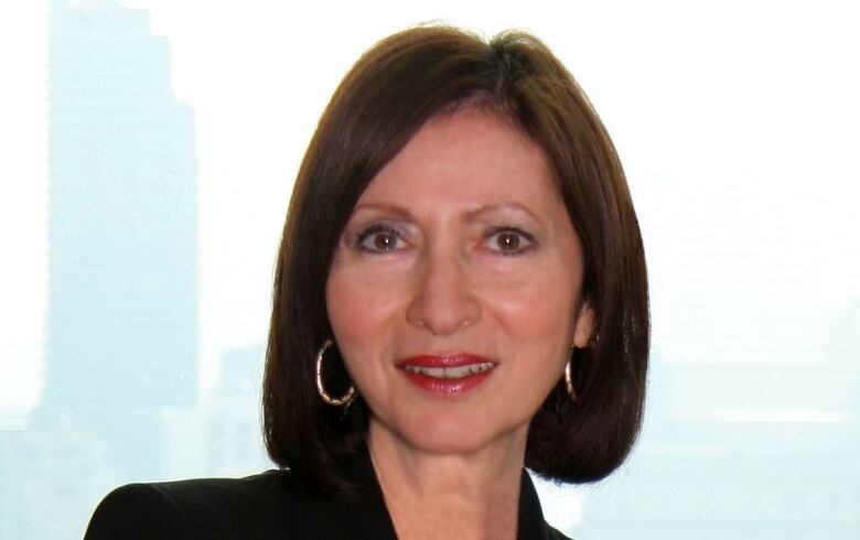 A smiling white woman with straight brown hair wearing a black blazer stands facing the camera with her arms crossed.