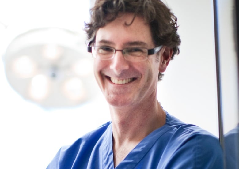 A smiling white man wearing blue scrubs with short, wavy brown hair and glasses leans against a wall, smiling at the camera.