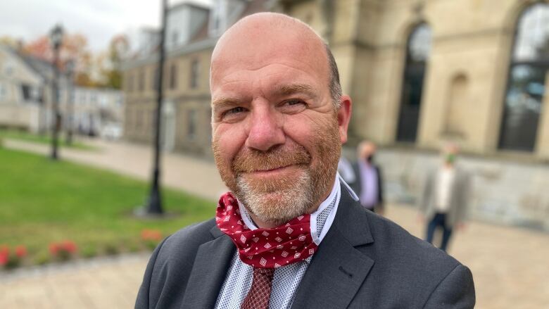 A man in a blue suit and red tie stands outside smiling at the camera.