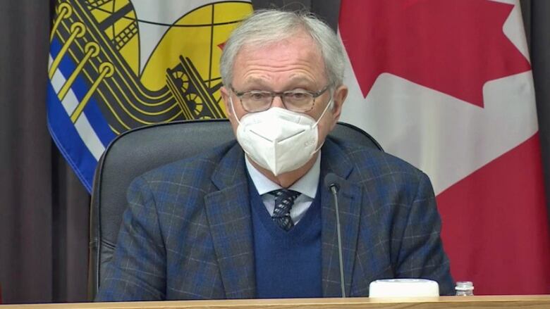 Man in suit sitting at table, wearing mask, Canada and New Brunswick flags in the background