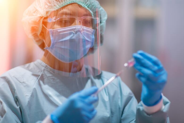 Woman in medical blue scrubs, face shield, mask and gloves holds a syringe.