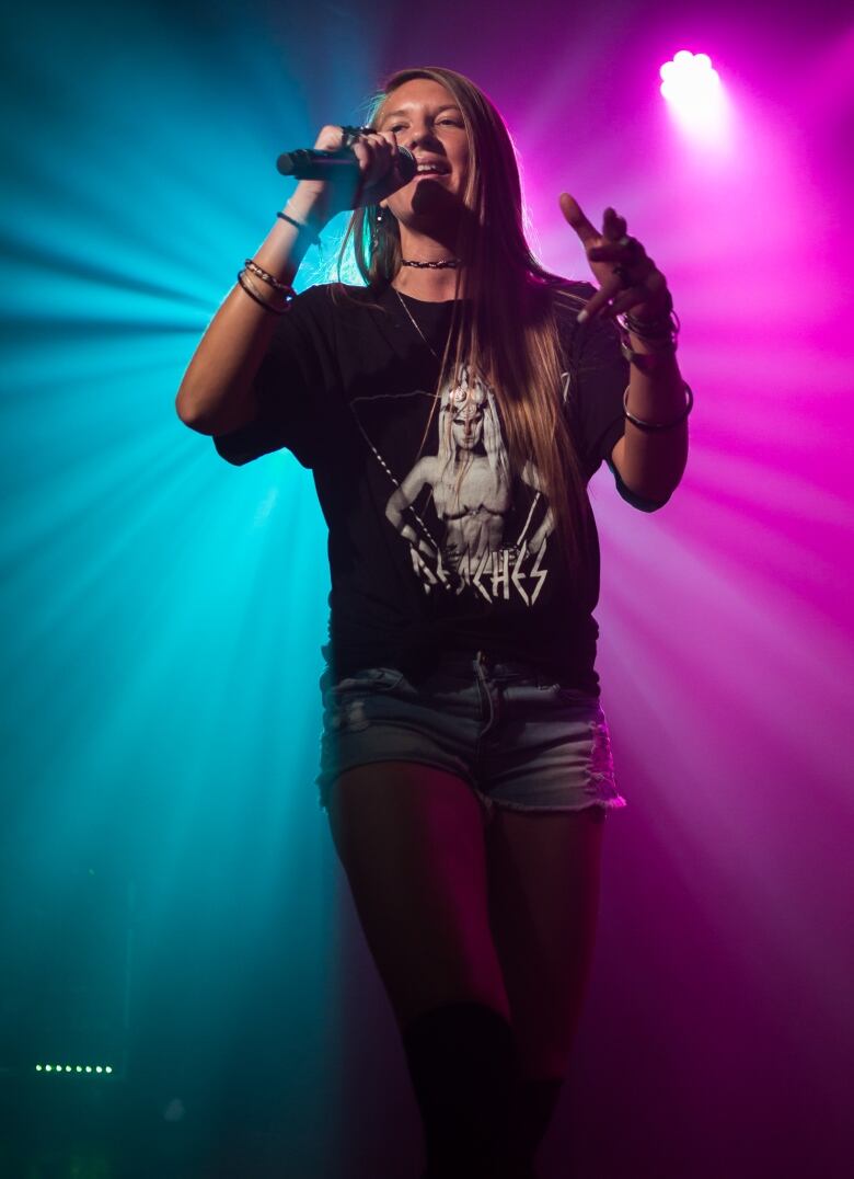 A white woman in a long black tee sings on stage in front of blue and purple lights