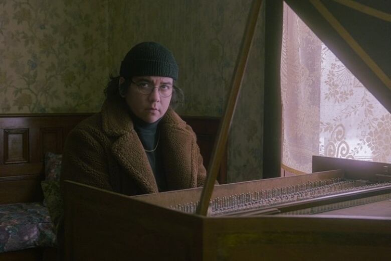 An Indigenous man with long brunette hair, glasses and a toque on sits at a piano