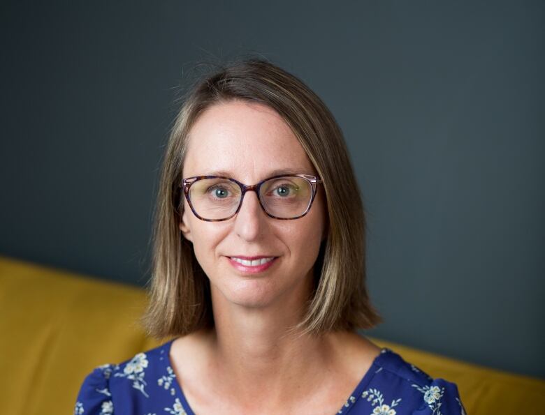 A woman with glasses and shoulder-length brown hair sits on a yellow couch.