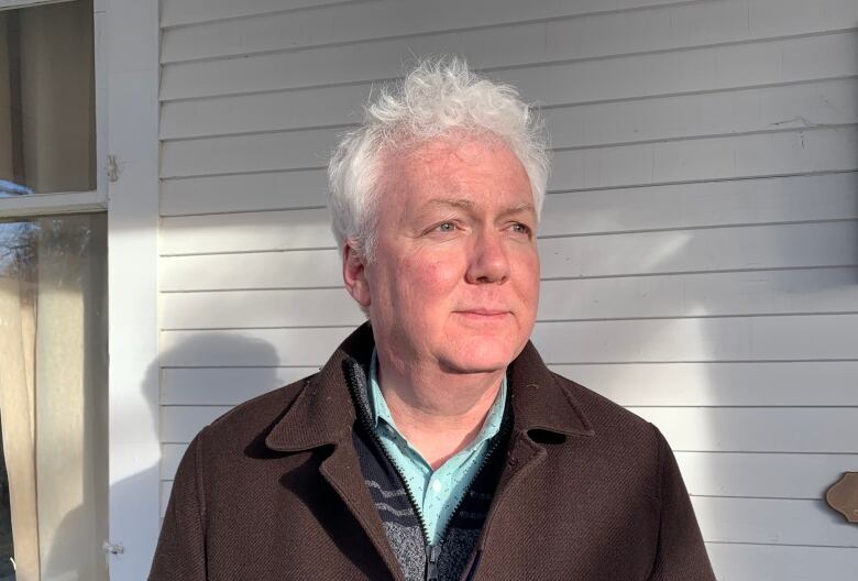 A man with white hair wearing a brown jacket stands in front of a barn.