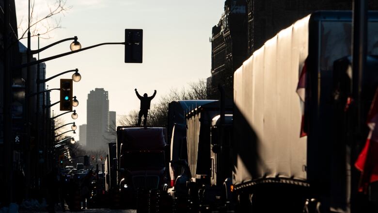 A man with his hands in the air is seen in silhouette while standing on top of one of several transport trucks parked on a downtown street.