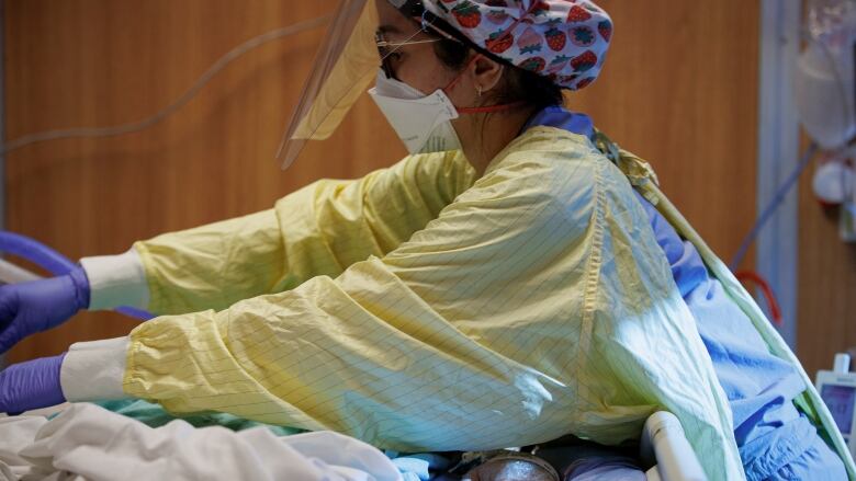 A woman in full PPE leans over a patient on a ventilator.