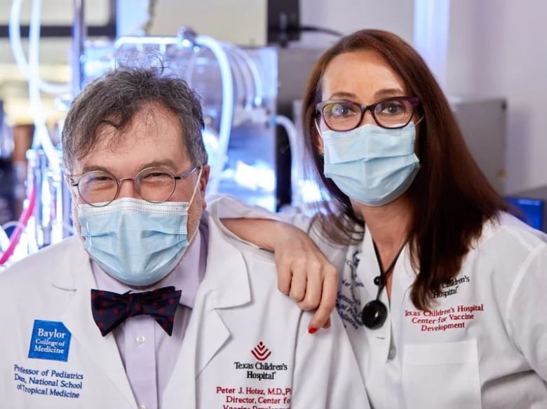 A man and woman doctor stand beside eachother. The woman, on the right, has her arm on the man's shoulder. Both are wearing white coats and blue medical masks. 