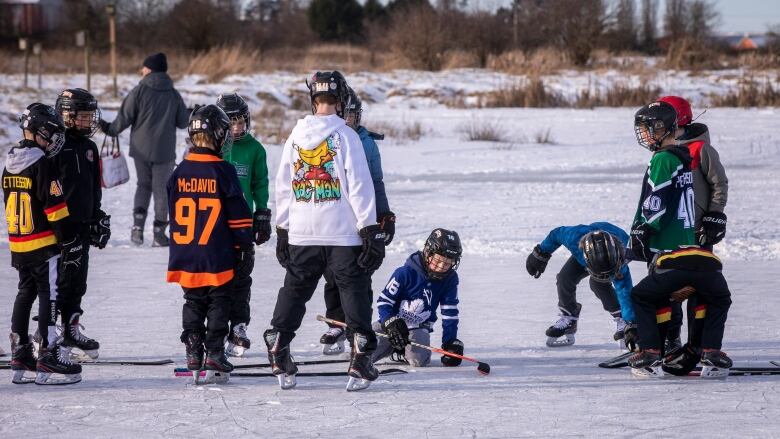 A group of children hang out in hockey gear on the ice outside.