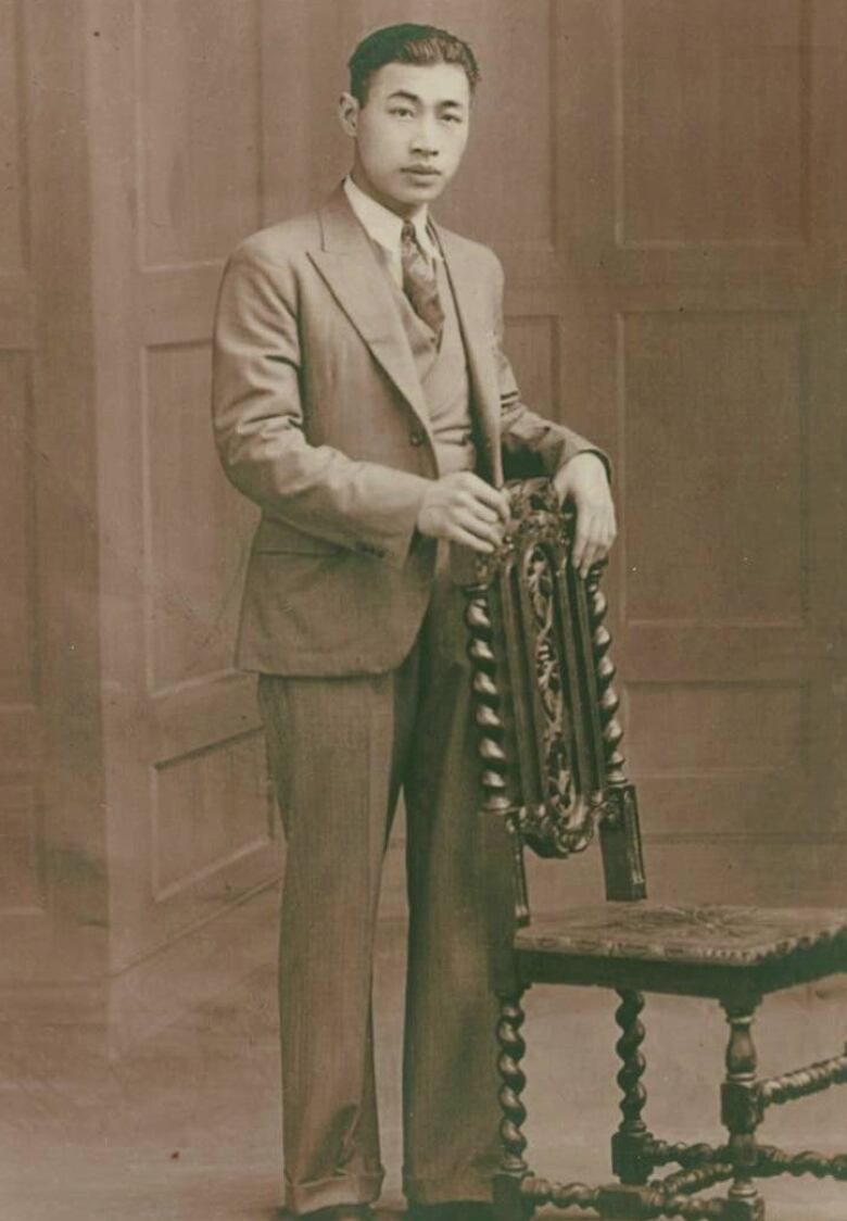 A black and white picture of a young Chinese man standing behind a chair. 