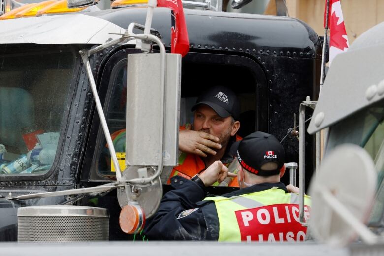 A police officer with a red 'POLICE LIASON' label on his back talks to someone in a truck in a city in winter.