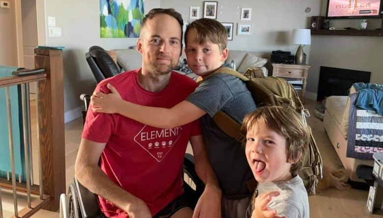 A man in a maroon T-shirt in a room at home with a child on his lap hugging him looks at the camera while one of his other children makes a face.