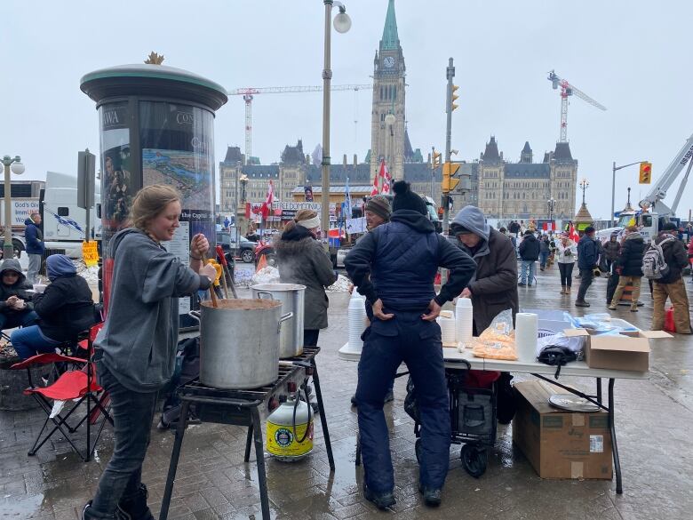 People stir and serve food in a square in front of a legislature in winter. There's a protest crowd around them.