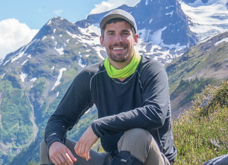 A man sits in a mountain meadow wearing a long-sleeved t-shirt and a baseball hat. He is smiling.