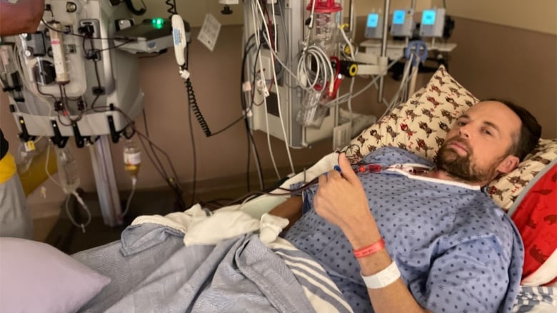 A man surrounded by hospital equipment lies in a hospital bed, looking at the camera.