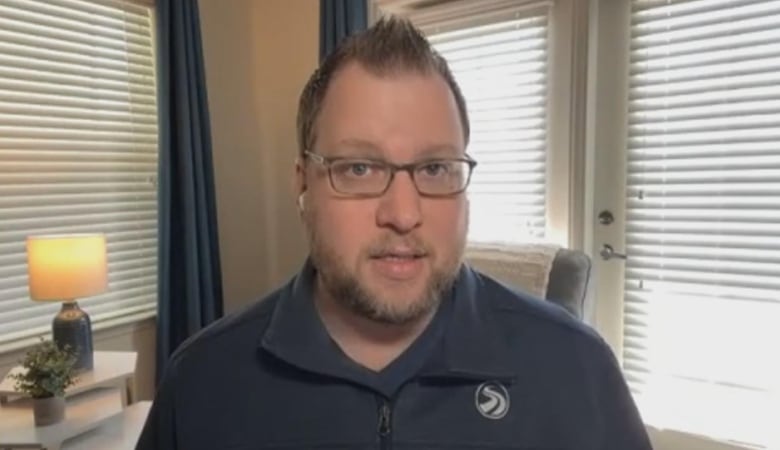 A portrait of a man with short brown hair, a beard and glasses, with closed blinds behind him, blocking out the sun.