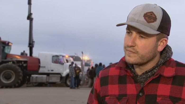 A while man in a red and black buffalo check jacket with a ball cap on. He's standing in front of a tractor and a truck.