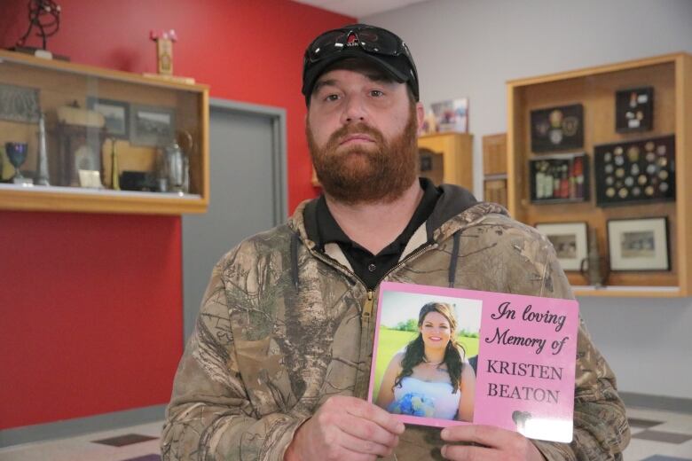 A man holds a small pink sign that reads, 'In loving memory of Kristen Beaton,' with a picture of a woman.