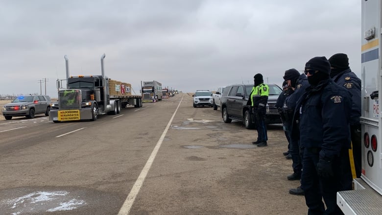 A line of semi trucks stretches down one side of a highway while R-C-M-P observe.