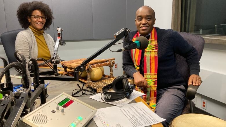 A Black woman with a fro sits in a radio booth with a Black man waring a black sweater