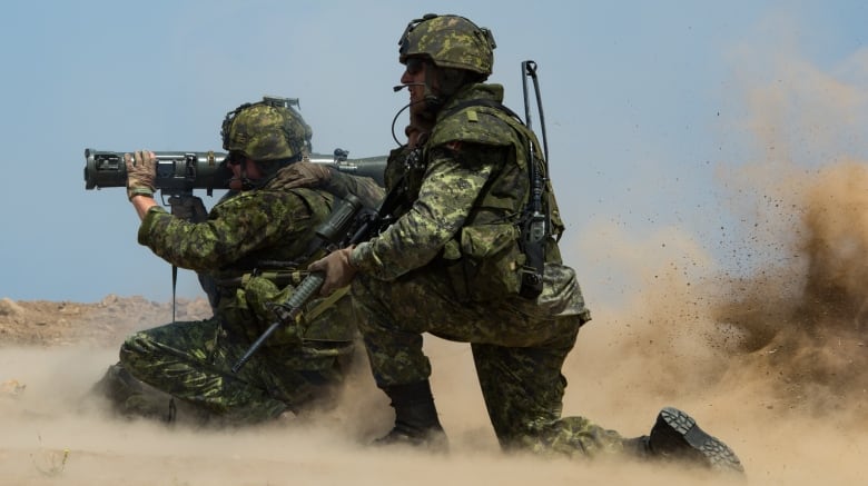 Two men in military camouflage kneel in an area of sand or lose dirt. One has a large cylindrical weapon on his shoulder. The other man, slightly behind him, holds an assault rifle.