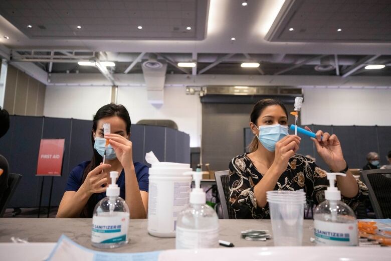 Two women hold syringes over small bottles at a table with plastic cups, hand sanitizer in a vaccine clinic