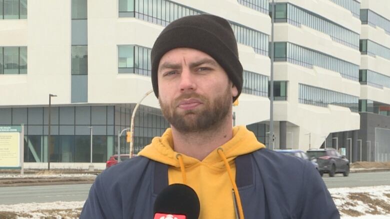 A young man in his 30s with a beard and a yellow hoodie, wearing a beanie. He's standing outdoors.