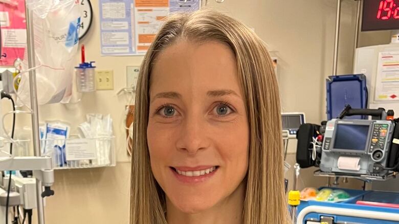 A doctor with long blonde hair stands in a hospital room with a stethoscope around her neck. 