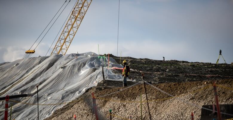 A worker wearing a yellow hard hat and a safety vest stands on a pile of dirt at a construction site near a crane.