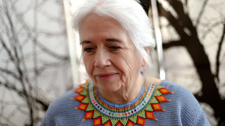 A woman with long whit hair looking at the ground a short distance ahead of her. She is wearing a periwinkle shirt with a beaded collar.