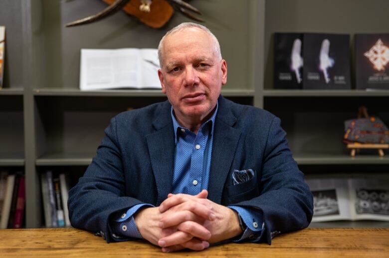 A man in a blue blazer sits with his hands folded at a wooden desk.