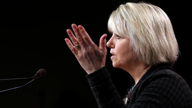 Dr. Bonnie Henry, a blonde woman with short hair, gestures at a podium.