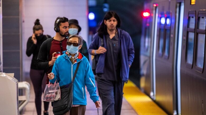 A group of people walk alongside a train in a subway. Some of them are wearing facemasks and others are not.