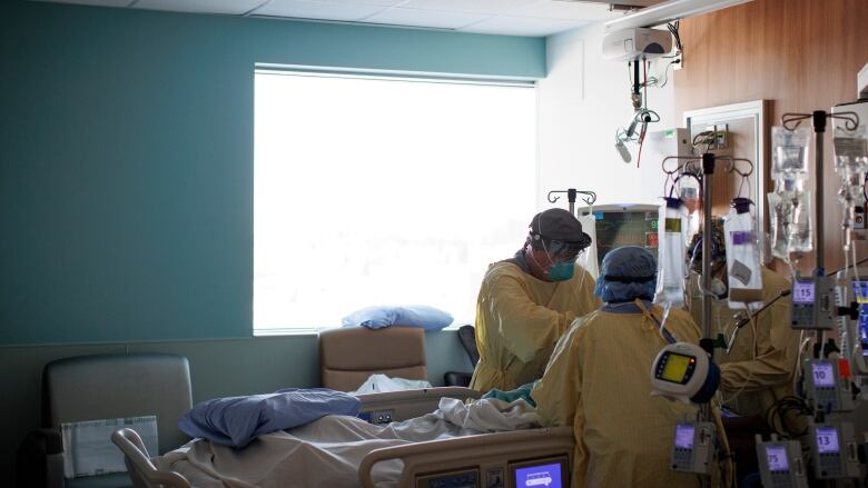 Three people in medical gowns, masks and caps stand around the head of a hospital bed, working on a patient. They are surrounded by medical equipment.