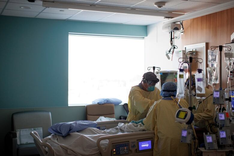 Three people in medical gowns, masks and caps stand around the head of a hospital bed, working on a patient. They are surrounded by medical equipment.