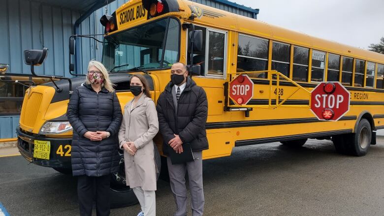 Two women and a man stand in front of a yellow school bus. 