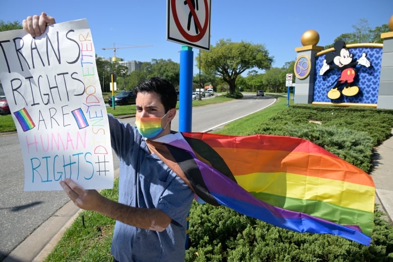 A man with a rainbow-coloured face mask and flag worn as a cape stands in front of a sign with Mickey Mouse on it, holding a sign that reads: Trans rights are human rights, Disney do better.