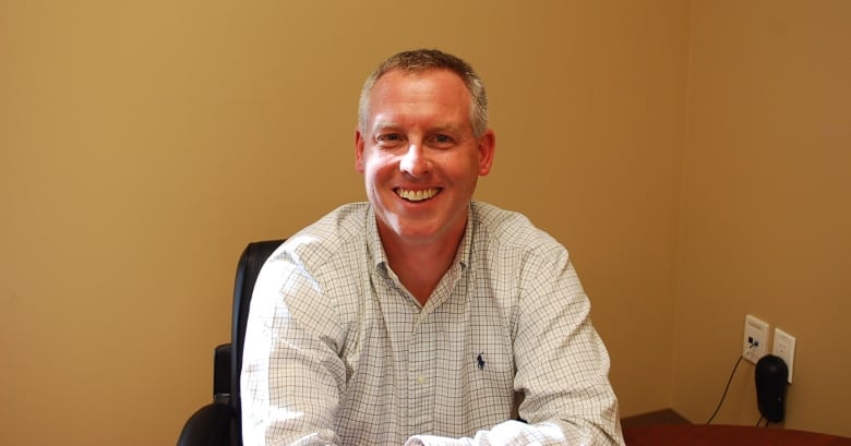 A man with light coloured hair and wearing a light colored checked shirt leans forward on a desk and smiles at the camera 