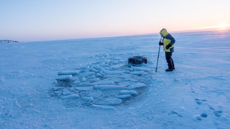 A person standing on sea ice looks at a big, partially frozen hole. 