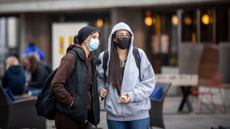 Two women wear masks and jackets in an outdoor environment.