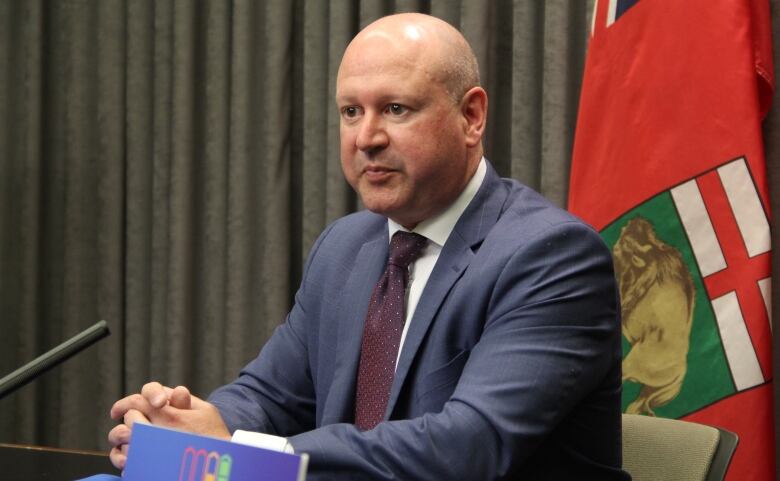 A man in a suit sits in front of a microphone at a desk, with Manitoba's flag behind him.