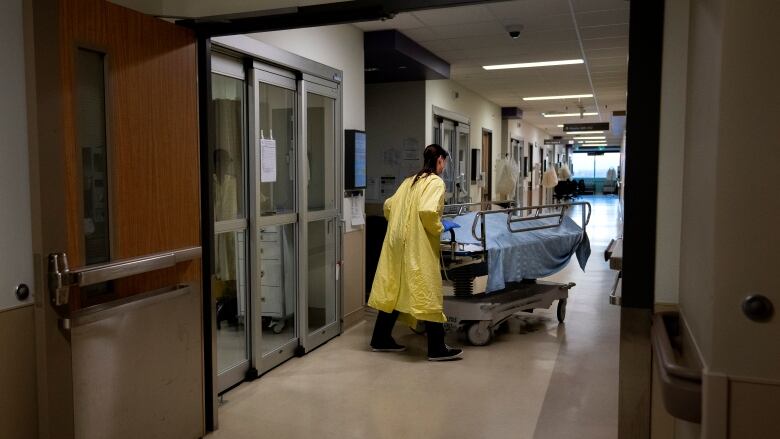 A health-care worker wearing a yellow medical gown transports a deceased COVID-19 patient on a stretcher, covered with a blue sheet, to the hospital morgue.