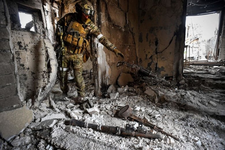 A soldier dressed in full military gear, including fatigues, a vest, a helmet and protective glasses, walks through a burnt-out room.