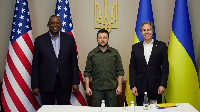 Three men, one in green military garb, the others in suits, stand before U.S. and   Ukrainian flags. 