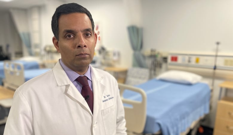  A man in a doctor's coat stands beside a hospital bed. His expression is sombre.