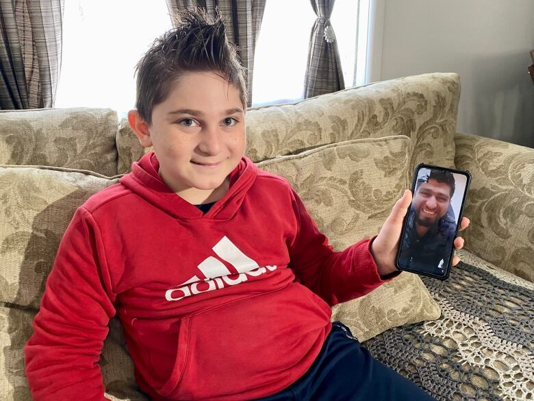 9-year-old boy with red sweatshirt and brown hair holds up cellphone with smiling face of his father.