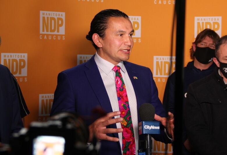 A man in a suit speaks at a podium, in front of a backdrop with the Manitoba NDP logo.